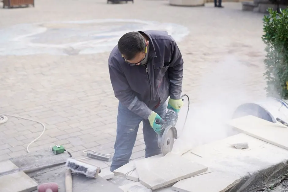 A construction worker uses a power saw to cut stone tiles outdoors, creating dust as he works.