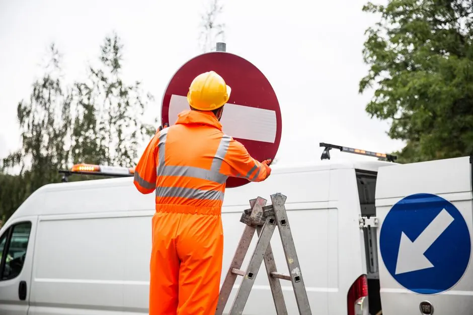 A road worker in orange uniform adjusting a traffic sign on a ladder by a van.